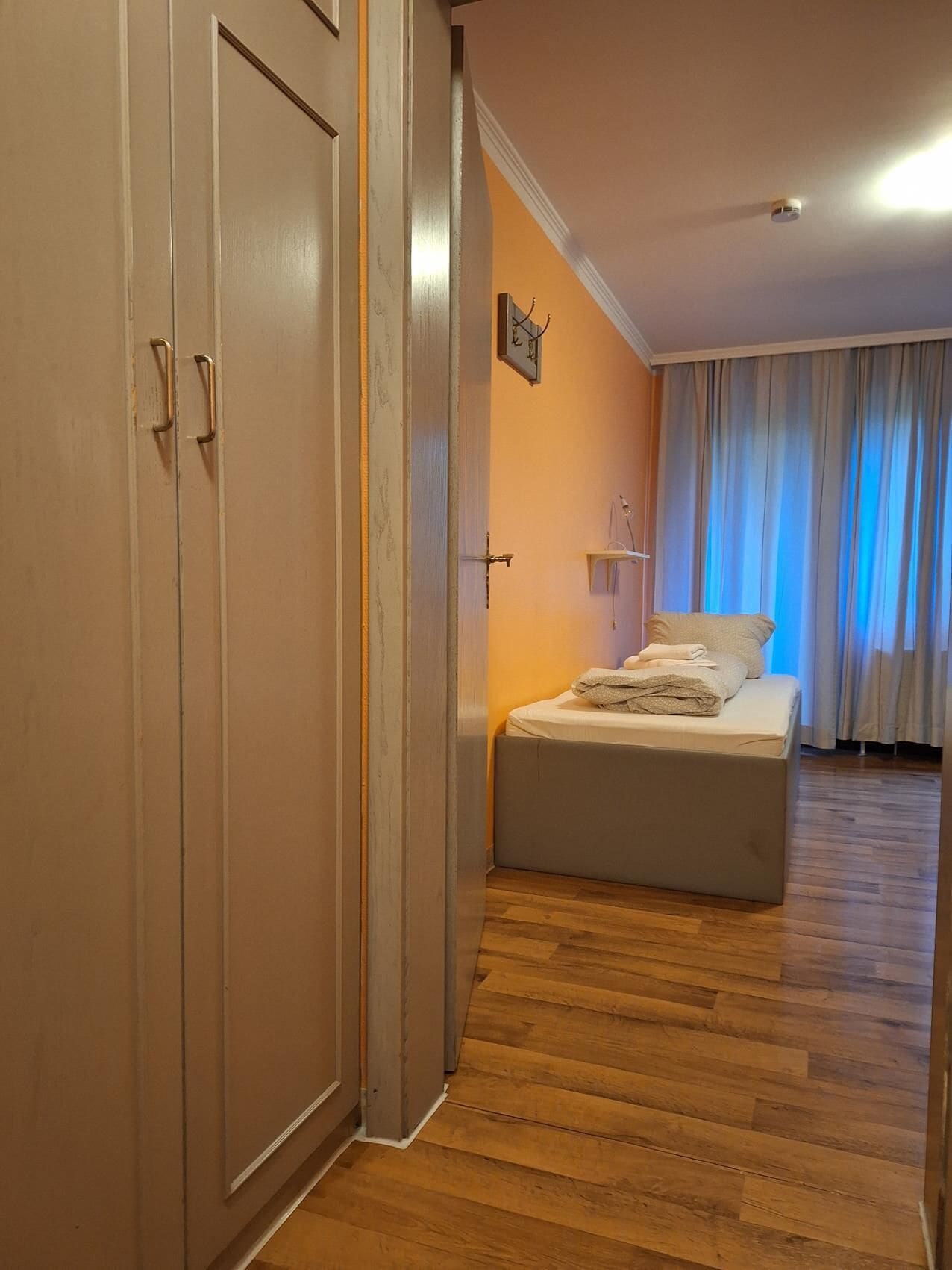 Un dormitorio con cama, suelo de madera, armario y ventana con cortinas.