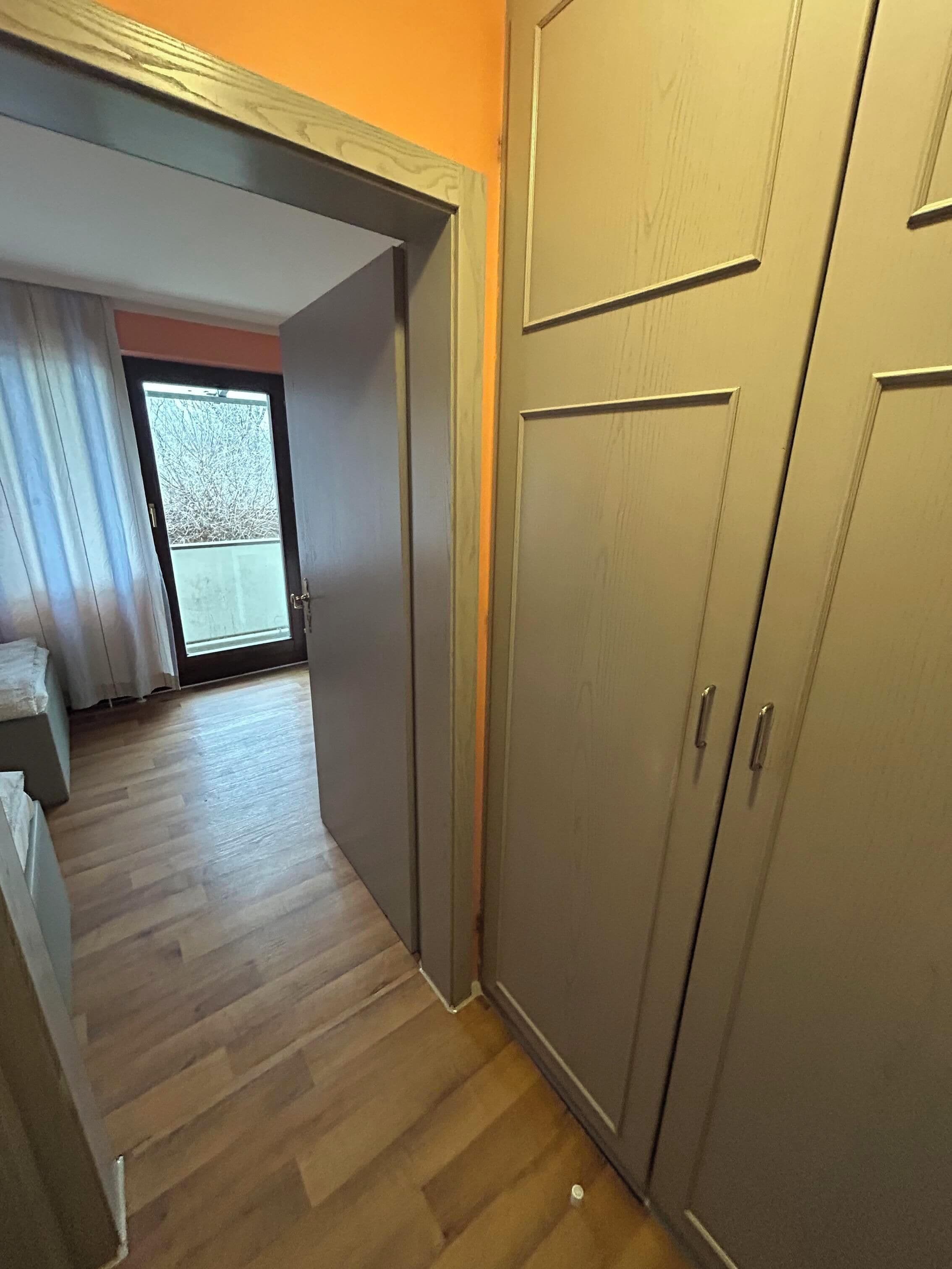A hallway with a wooden floor leading to a room with a large wardrobe and a door leading outside.