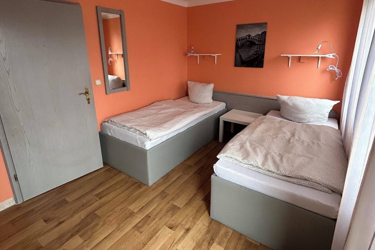 Une chambre de taille modeste, équipée de deux lits simples, d’une petite table, d’un miroir et d’une étagère sur laquelle est posée une image.