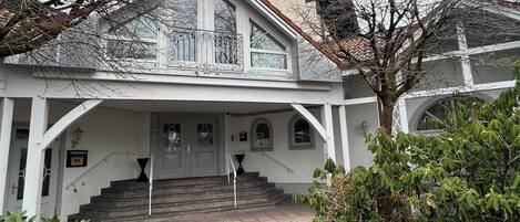 Ein zweistöckiges Gebäude mit einer symmetrischen Fassade, einem zentralen Eingang mit Treppe und einem kleinen Balkon mit aufwendiger Geländerarbeiten.