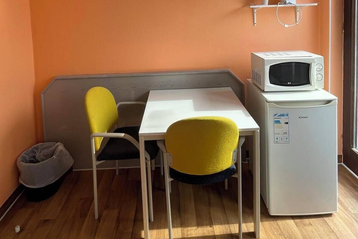 Une petite cuisine équipée d’un four à micro-ondes, d’une table blanche, de deux chaises, d’un banc et d’une étagère sur laquelle se trouvent une tasse et un téléphone.