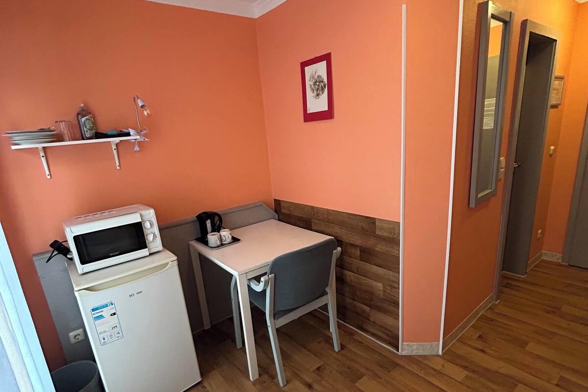 Une cuisine compacte équipée d’un four à micro-ondes, d’un petit réfrigérateur et d’une table avec une chaise. Une étagère fixée au mur est garnie de divers objets, et un tableau encadré y est également accroché.