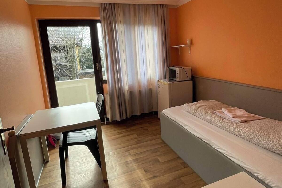 Une petite pièce bien éclairée, avec un lit, un bureau et une fenêtre avec des rideaux.