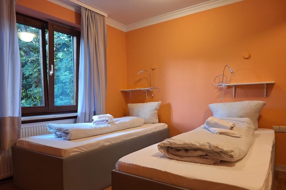 Une chambre avec deux lits simples, chacun doté d’une table de chevet et d’une lampe. Il y a une fenêtre avec des rideaux, et les murs sont peints en orange.
