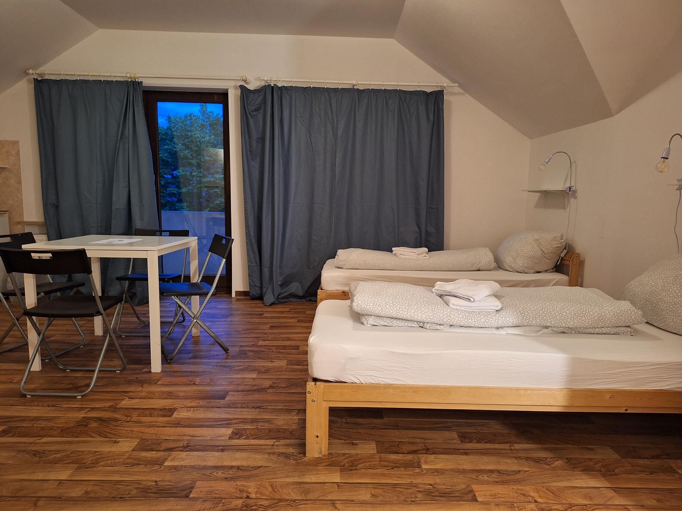 Ein Zimmer mit Holzboden, einem Bett mit weißer Bettwäsche, einem kleinen Tisch mit Stühlen und einem Fenster mit Vorhängen.