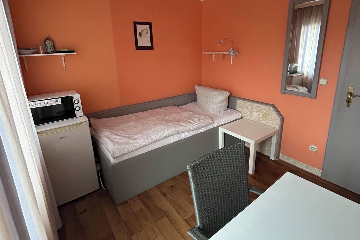 Une petite chambre comprenant un lit, une petite table, une chaise, un four à micro-ondes et une étagère avec quelques objets.