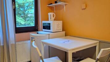 Una habitación compacta con una pequeña mesa y dos sillas, una ventana que ofrece vistas a árboles, un radiador y un microondas en un estante.