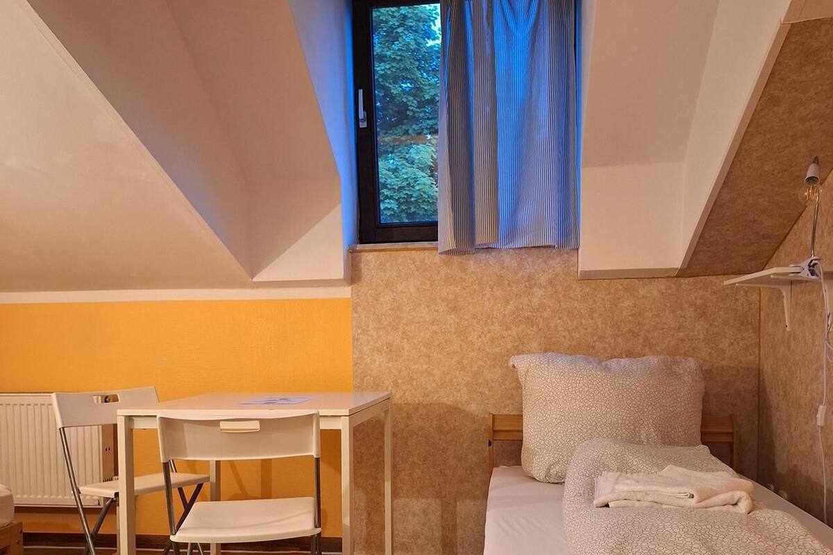 Une petite pièce bien éclairée, avec un lit, un bureau et une chaise. On y trouve une fenêtre avec des rideaux, un radiateur et un plancher en bois.