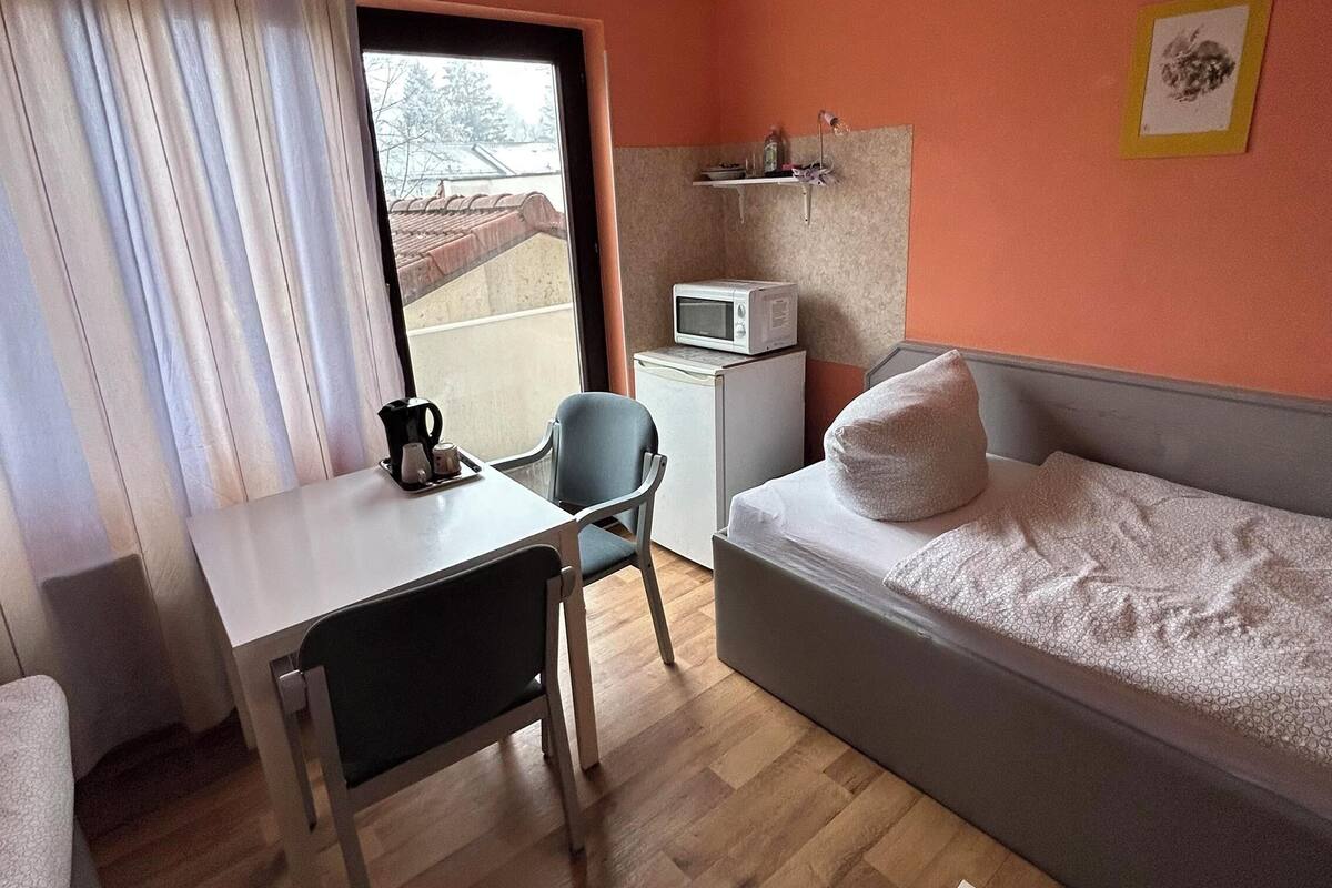 Une chambre d’hôtel de taille réduite, comprenant un lit, un bureau, une chaise, un four à micro-ondes et une fenêtre avec des rideaux.