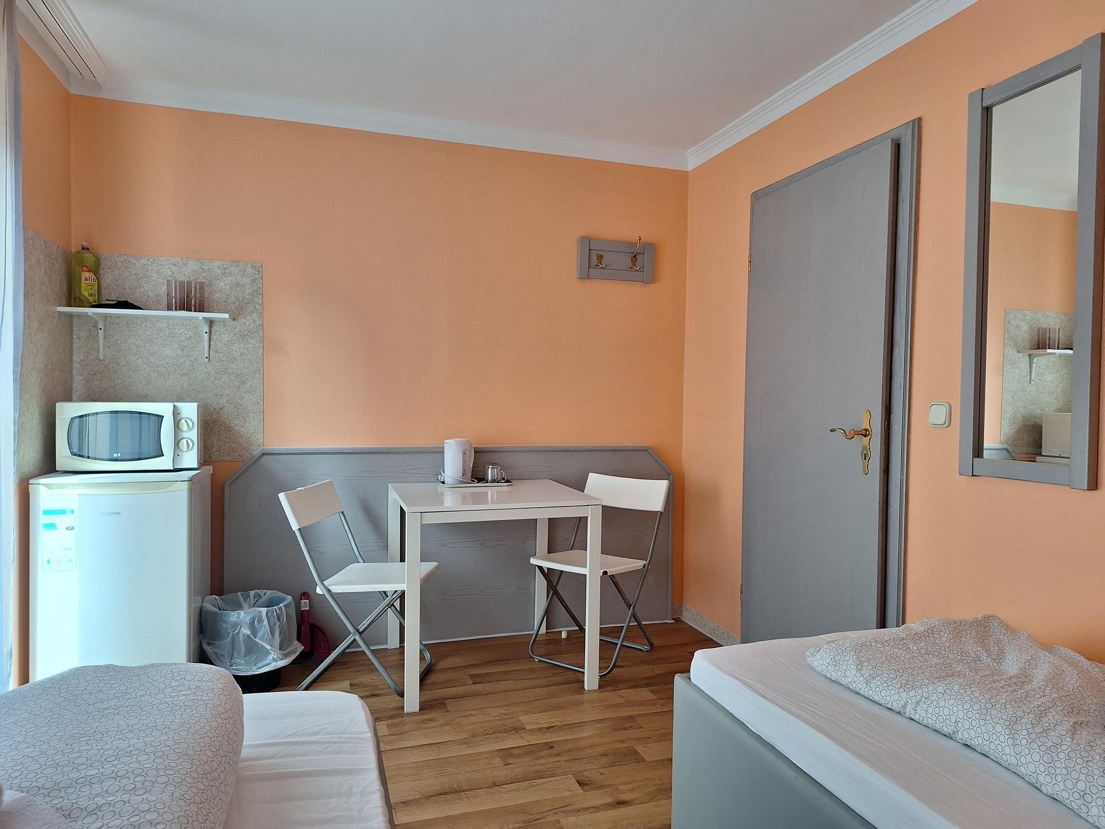 A small, neatly arranged room with a bed, a desk with a chair, a microwave, and a shelf with items.