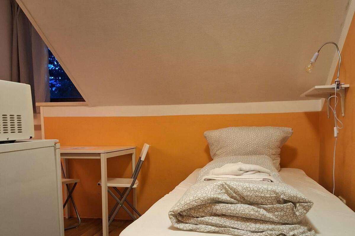 Un lit simple avec une couverture à motifs, un bureau avec une chaise et une petite table avec une lampe.