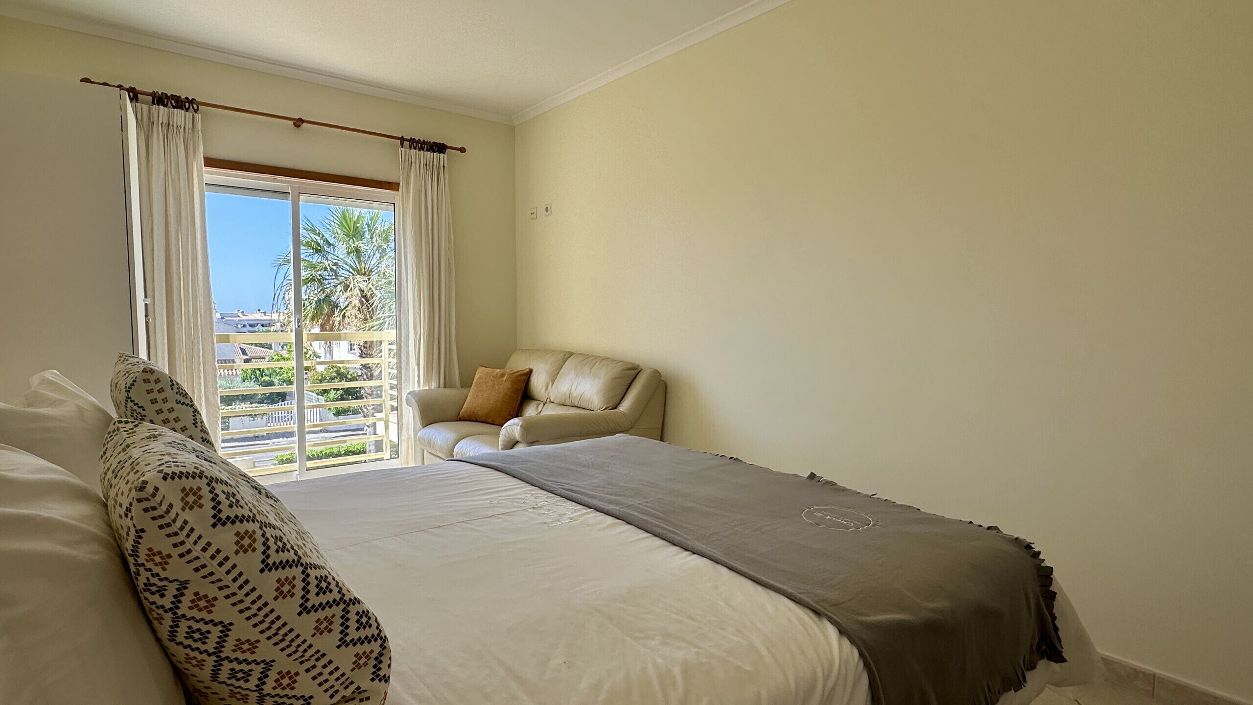Quarto com cama, uma cadeira e vista para palmeiras pela janela.
