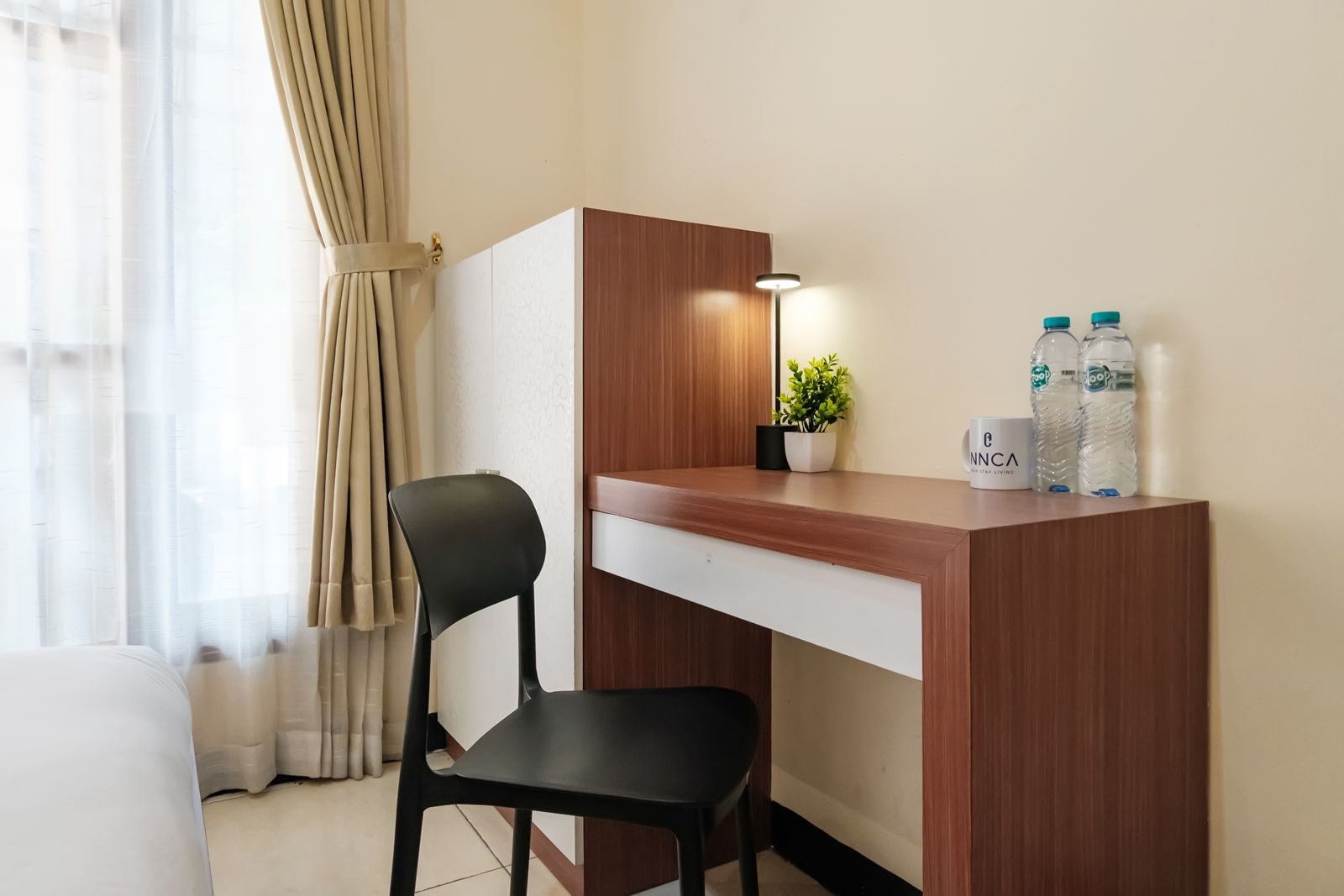 Camera d'albergo con scrivania, sedia, pianta in vaso, bottiglie d'acqua e una finestra con tende.