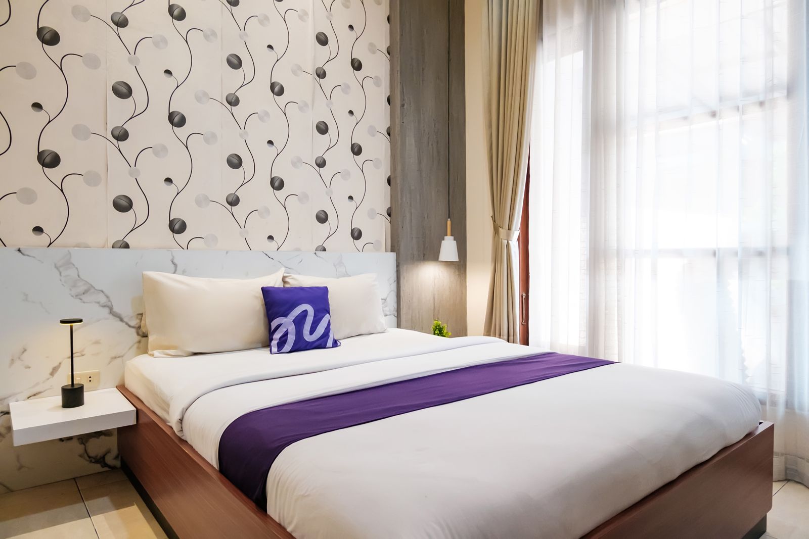 Une chambre à coucher moderne avec un grand lit, une table de chevet, une lampe et une vue sur l’extérieur à travers des rideaux légers.