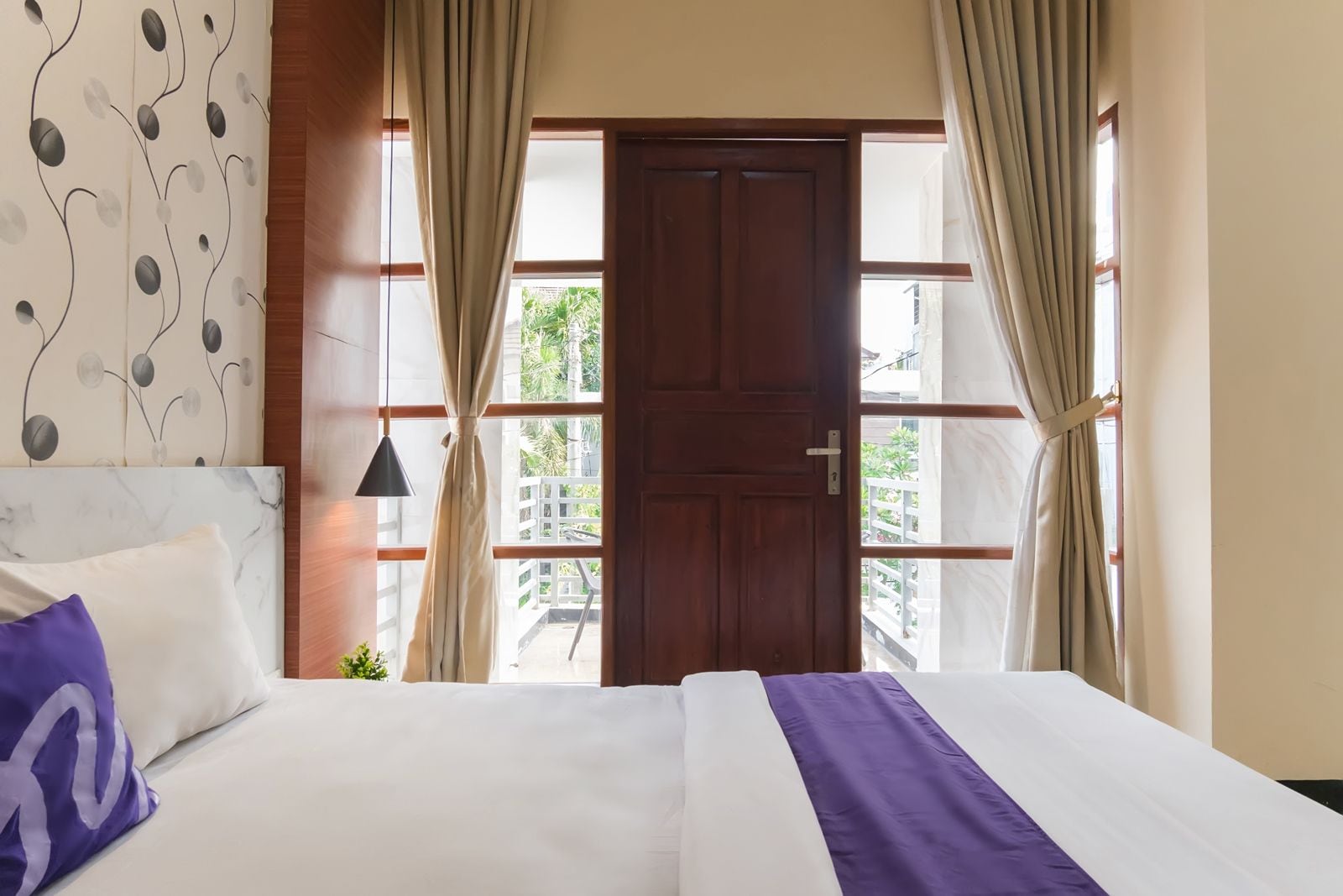 A hotel room with a bed, curtains, a door, and a view of the outside.