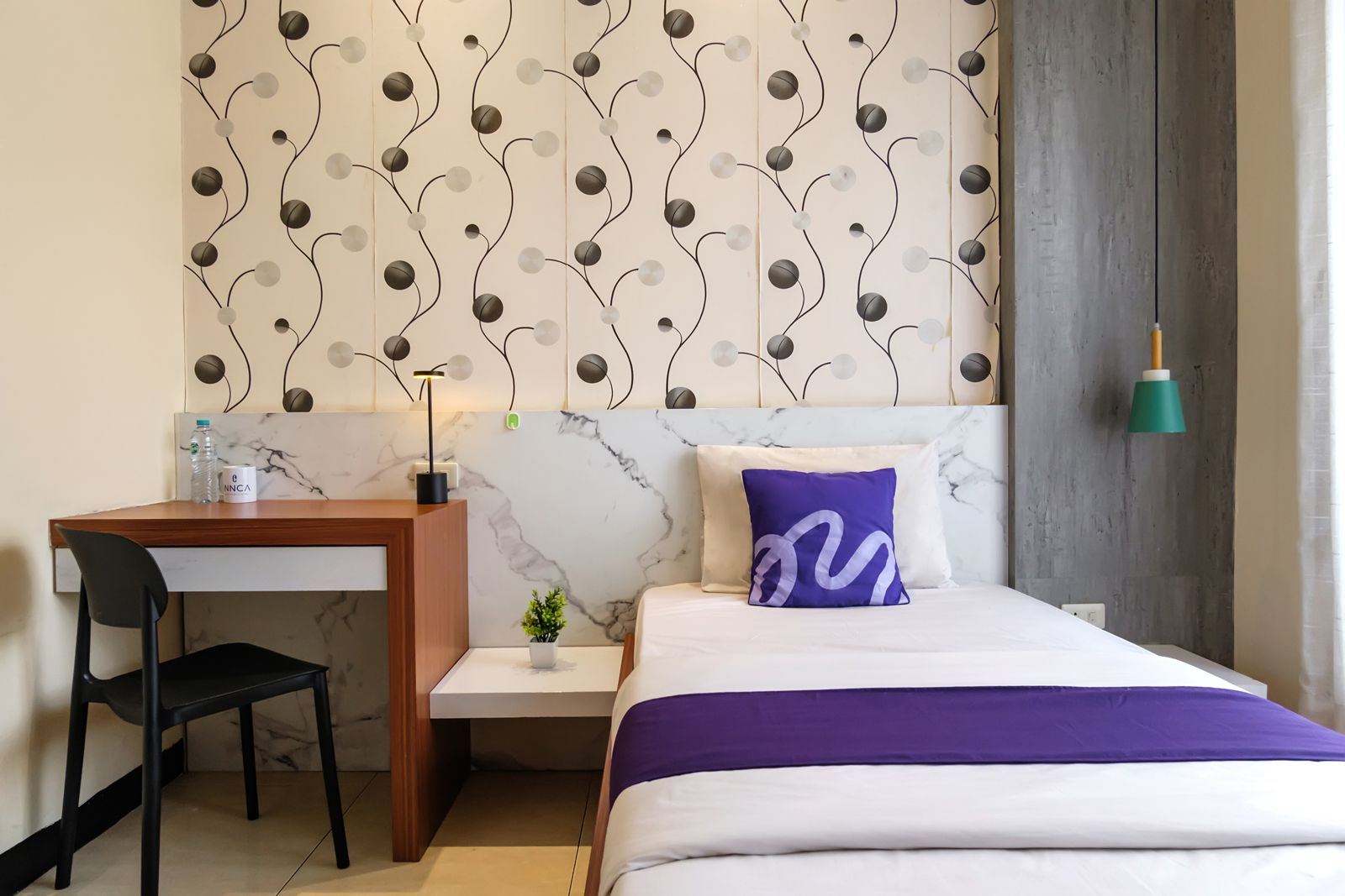 Een hotelkamer met een bed, een bureau, een stoel, een kleine plant en een muur met een decoratief patroon.