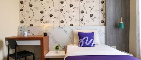 A hotel room with a bed, a desk, a chair, a small plant, and a wall with a decorative pattern.