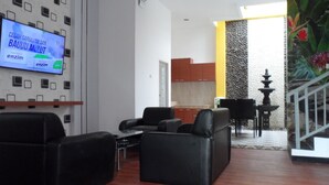 Un salon moderne avec des canapés en cuir noir, un téléviseur à écran plat fixé au mur et un coin cuisine visible à travers une porte ouverte.
