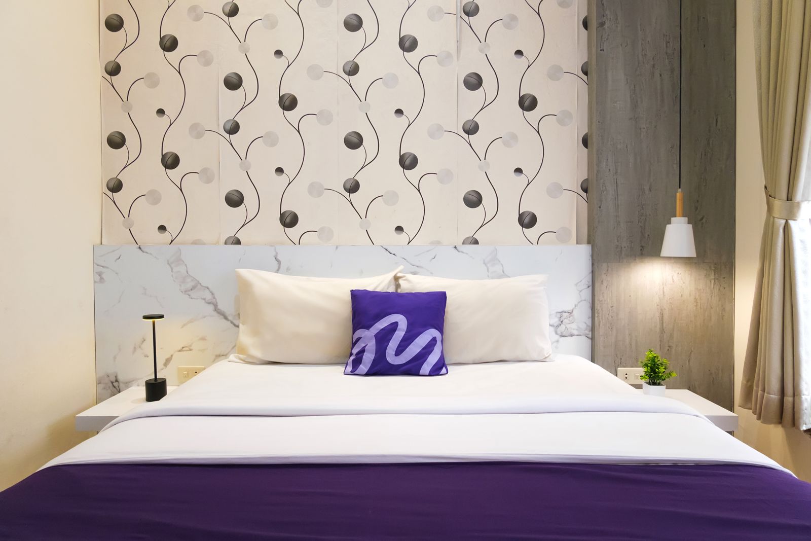 Ein Hotelzimmer mit einem Kopfende mit Marmoroptik, einem Bett mit weißer Bettwäsche und einer violetten Tagesdecke sowie einer Wand mit einem dekorativen Muster.