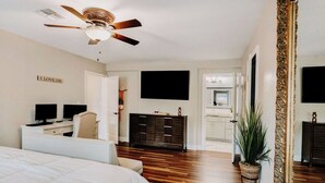 Un dormitorio con cama, escritorio con dos monitores, cómoda oscura, ventilador de techo, televisor y un espejo grande.