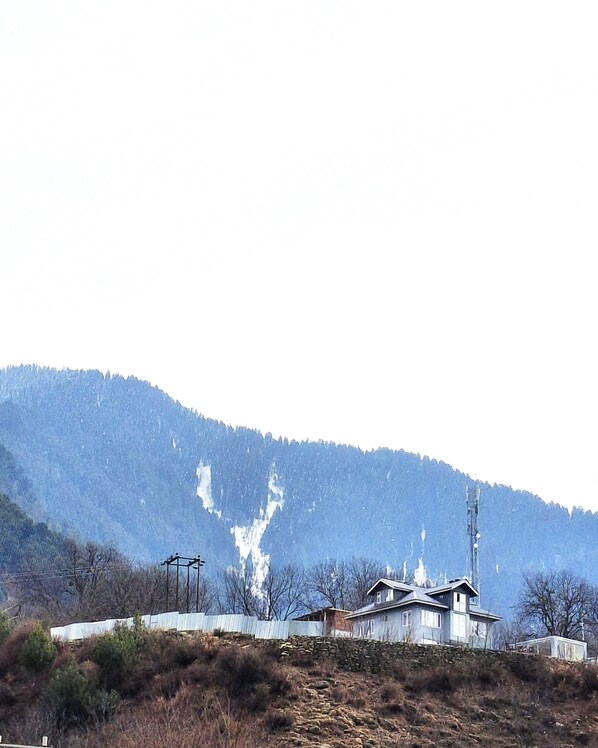Un edificio de dos pisos con techo inclinado, ubicado en una ladera con montañas al fondo.