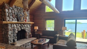 L’intérieur chaleureux d’un chalet avec une cheminée en pierre, des poutres en bois, un mur en pierre, une grande fenêtre donnant sur un lac et un coin salon confortable.
