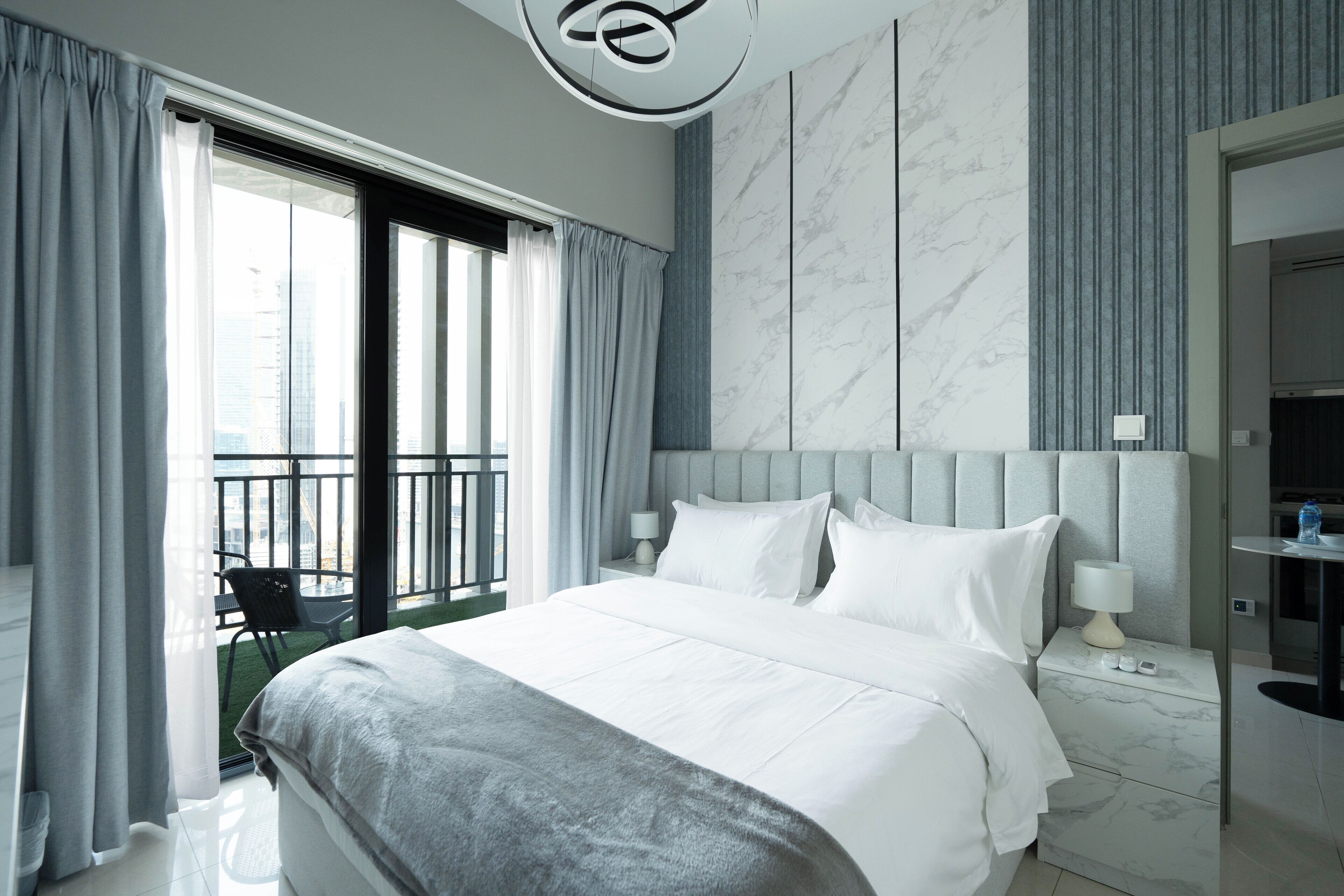 A modern hotel room with a large bed, a nightstand, a lamp, and a view of the city through a balcony.