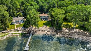 An aerial view of a lakeside property with a dock extending into the water, surrounded by dense forest and a large house with a well-maintained lawn.