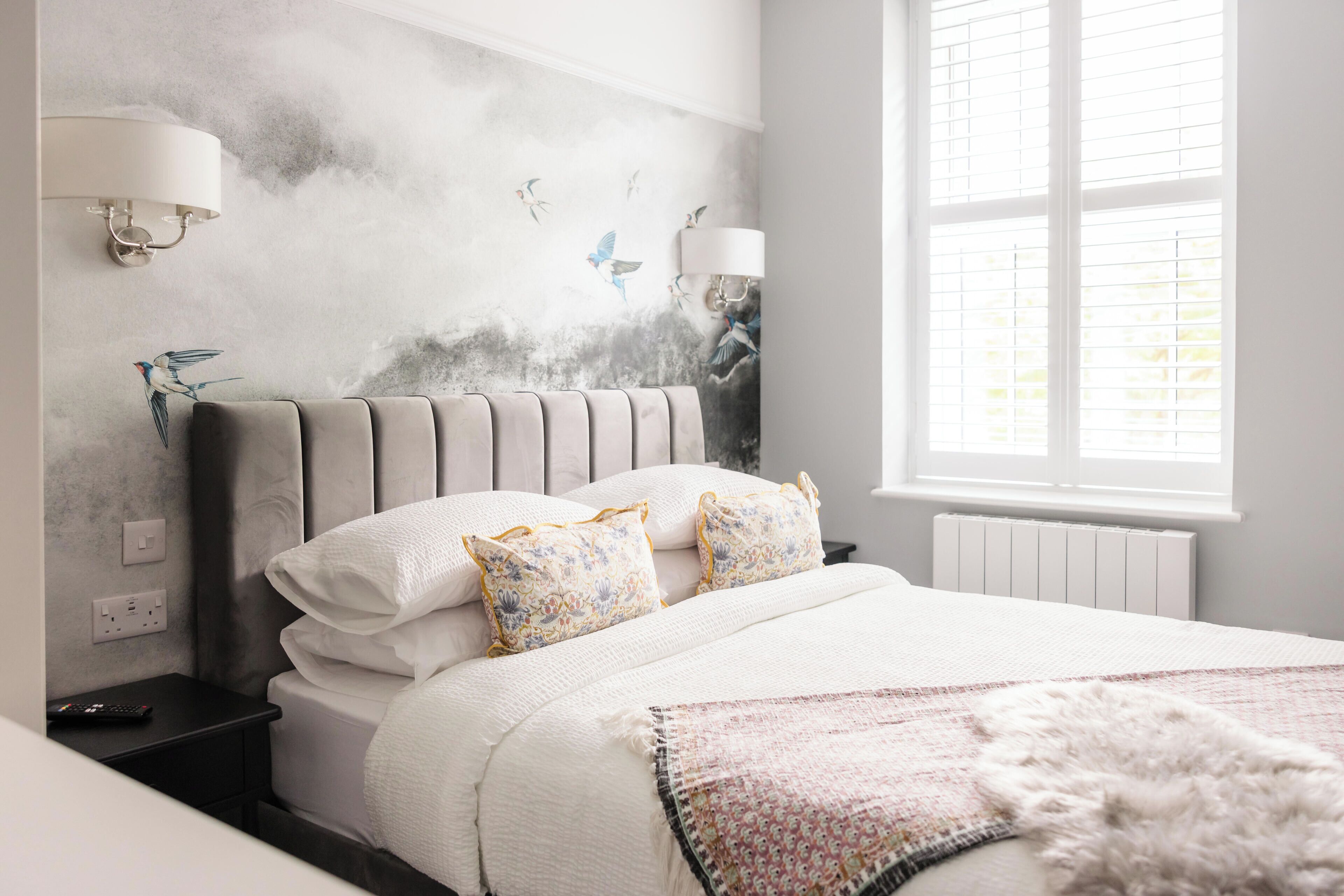 Un dormitorio con cabecera tapizada en gris, ropa de cama blanca y un mural decorativo en la pared.