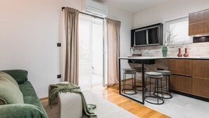 Un espacio habitable compacto con sofá, una otomana pequeña, zona de cocina con televisor, taburetes y salpicadero de ladrillo.