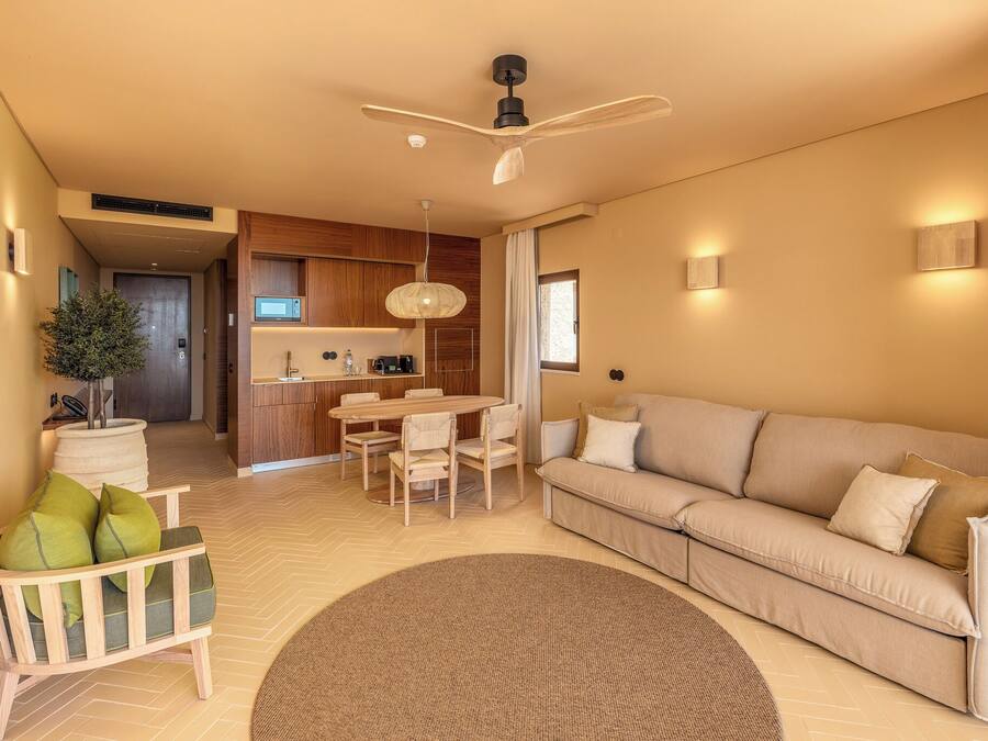Ein modernes Wohnzimmer mit einem beigefarbenen Sofa, einem Holzstuhl mit Kissen, einem Esstisch mit Stühlen, einem Deckenventilator und einem Küchenbereich im Hintergrund.