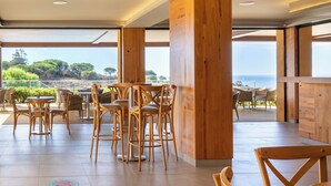 An outdoor dining area with wooden chairs and tables, large wooden pillars, and a view of the sea and trees.