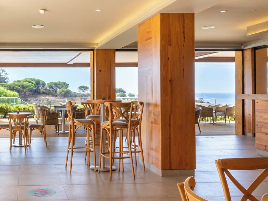 Un área de comedor al aire libre con sillas y mesas de madera, grandes pilares de madera y vistas al mar y a los árboles.
