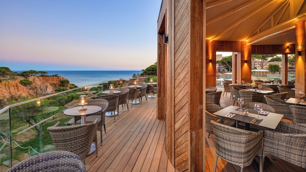 A wooden deck with wicker furniture and tables, overlooking a coastal landscape with cliffs and the sea.