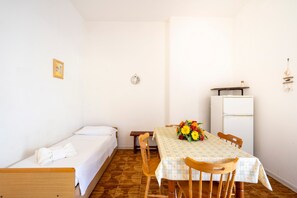 A small, clean room with a single bed, a dining table with chairs, a refrigerator, and a wall-mounted light fixture.