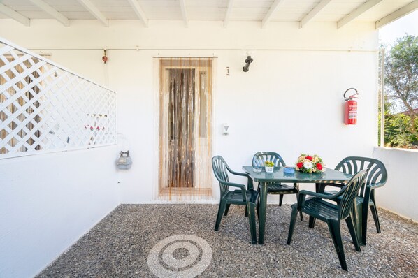 Eine überdachte Terrasse mit Tisch und Stühlen, einem Feuerlöscher und einer weißen Wand mit einer Tür.