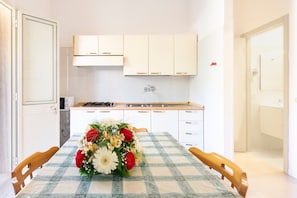 Une cuisine de taille modeste, avec des meubles blancs, une cuisinière à gaz et une nappe à carreaux agrémentée d’un centre de table floral.
