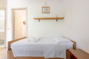 Une chambre avec un lit en bois, du linge de lit blanc, un banc en bois, une étagère fixée au mur et une porte donnant sur une autre pièce.