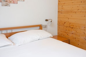 A bedroom with a wooden headboard, white bedding, a bedside table, and a wall-mounted light fixture.