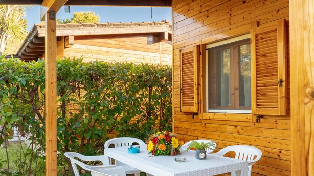 A wooden cabin with a covered patio, white plastic chairs, a table with a vase of flowers, and a window with shutters.