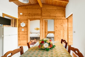 A wooden interior with a dining table set for four, a refrigerator, and a bed in the background.
