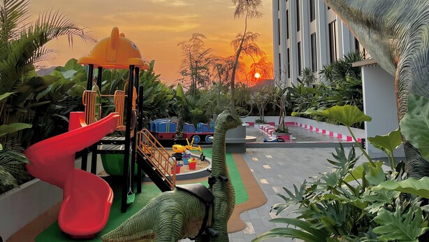 Ein Kinderspielbereich mit Rutsche und Klettergerüsten, umgeben von Grünflächen und einer Dinosaurierstatue.