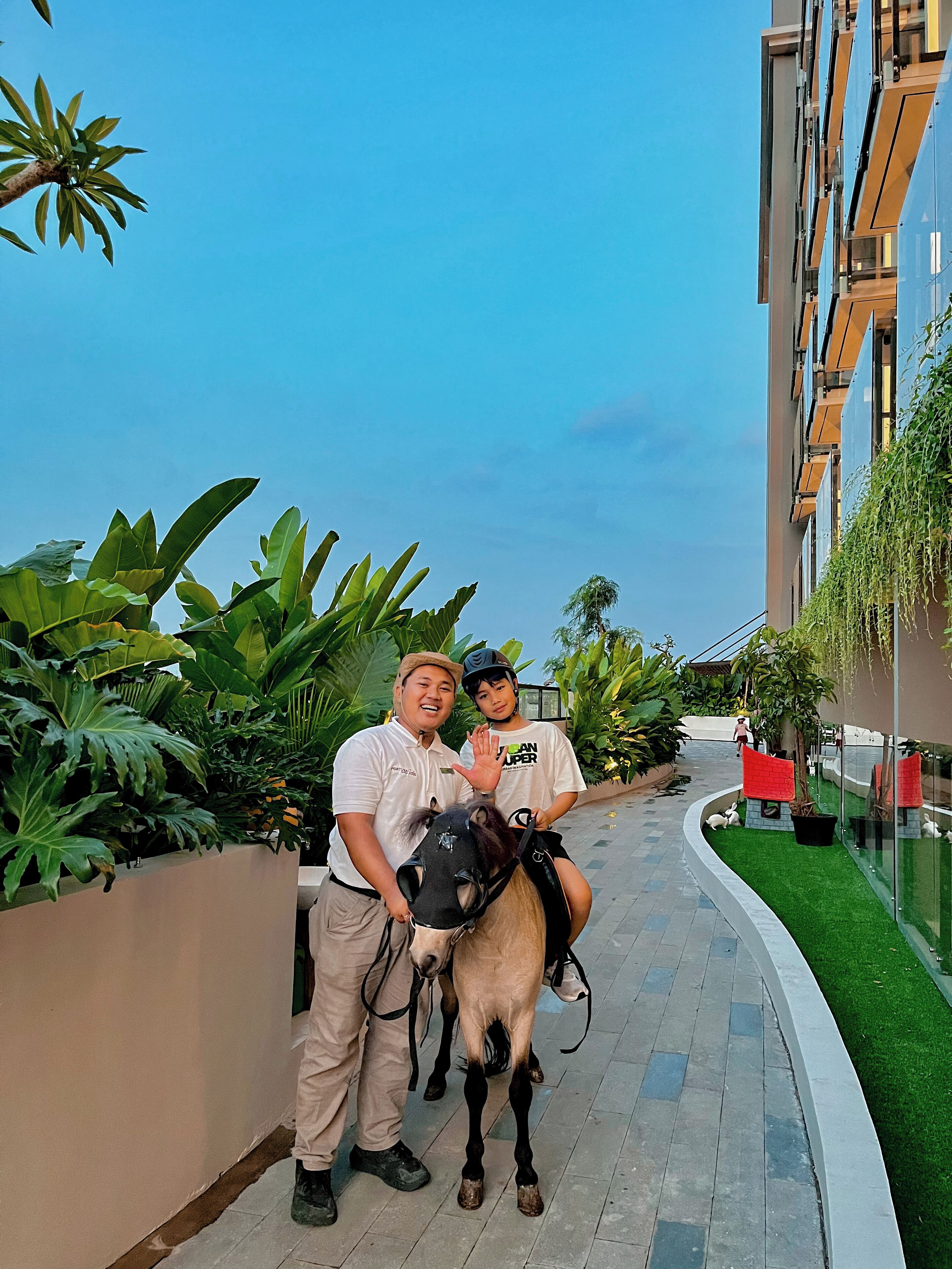 A man and a child on a horse in an outdoor area with a building and greenery in the background.