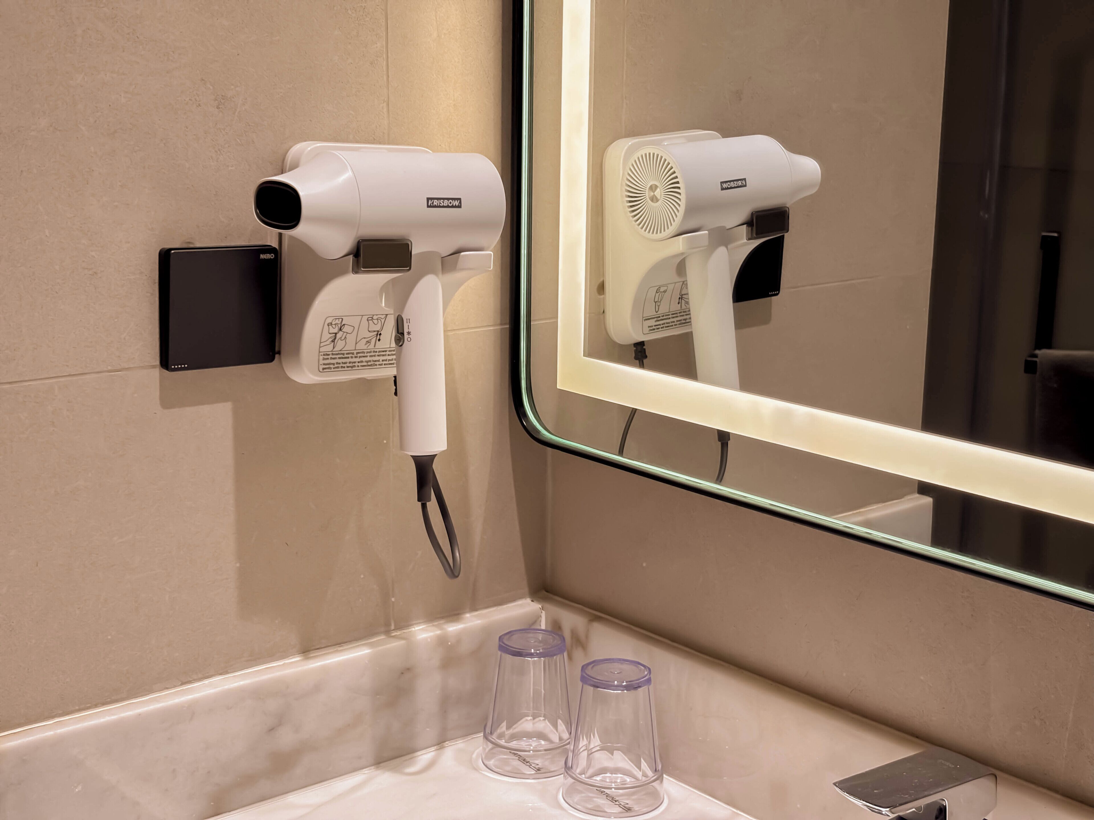 A bathroom with a hairdryer mounted on the wall, a mirror, and two empty glasses on the counter.