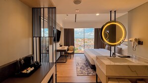 Ein modernes Hotelzimmer mit Bett, Schreibtisch, Sessel, Fernseher, einem großen Spiegel und Blick auf die Stadt.