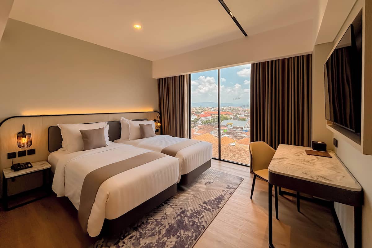 Habitación de hotel con dos camas, un escritorio, una silla, un televisor y un ventanal grande con vista a la ciudad.