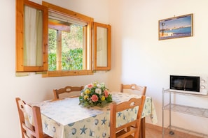 A dining area with a table set for two, a flower centerpiece, wooden chairs, a window with open shutters, and a framed picture on the wall.