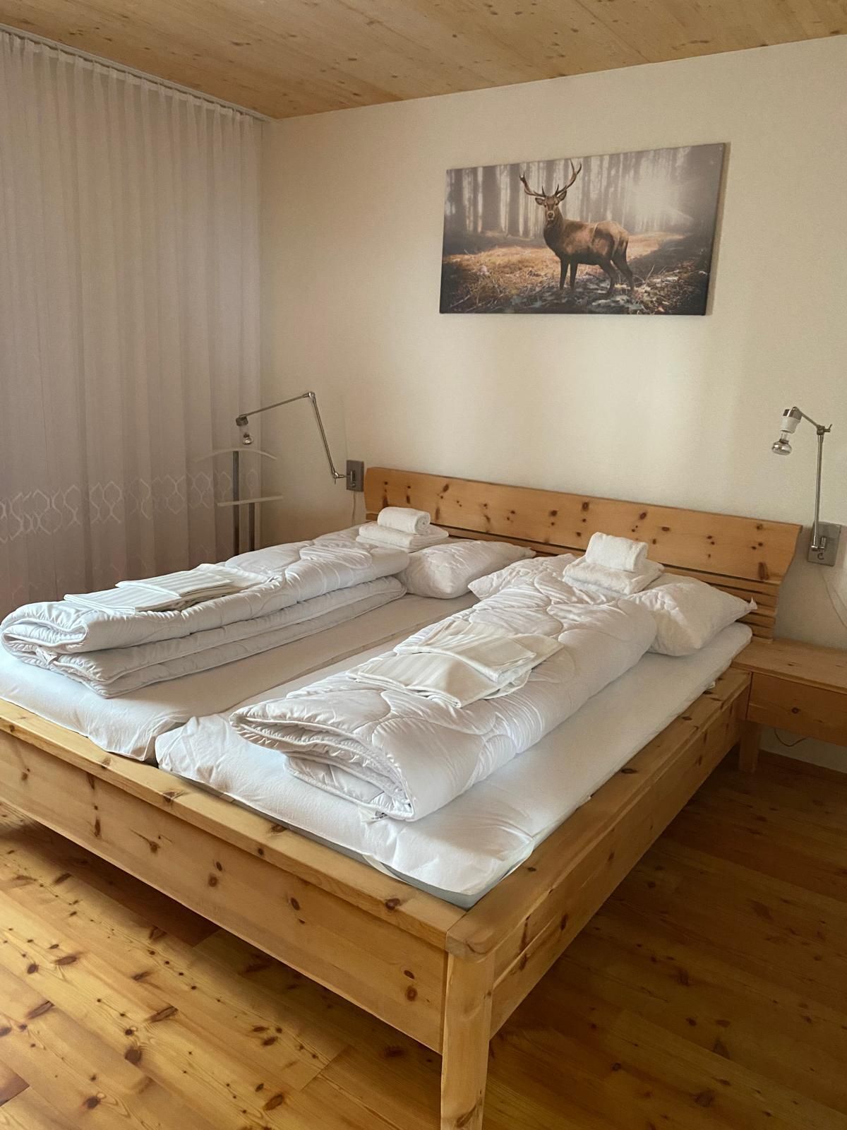 A wooden bed with white bedding, two bedside lamps, and a forest-themed painting on the wall.
