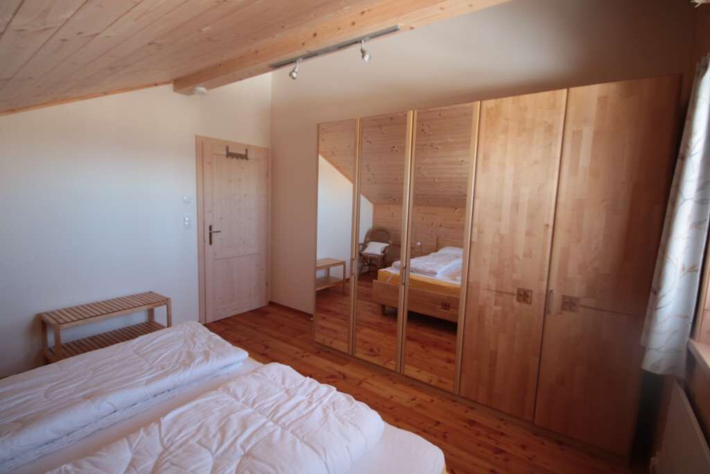 A bedroom with a wooden floor, a bed with white bedding, a wooden wardrobe, and a small wooden bench.