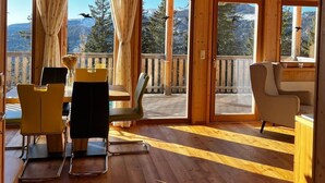 Een houten chalet van binnen, met een eethoek, stoelen en uitzicht op een besneeuwd berglandschap door grote ramen.